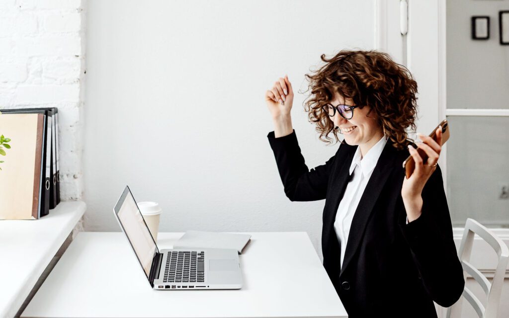 Junge und moderne Frau in Business-Outfit jubelt am Bürotisch.