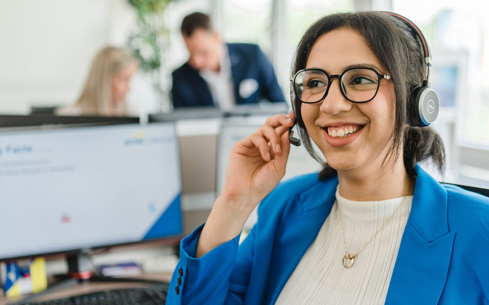 Freundliche, junge Mitarbeiterin in moderner Business-Kleidung und mit Headset sitzt am Bürotisch.