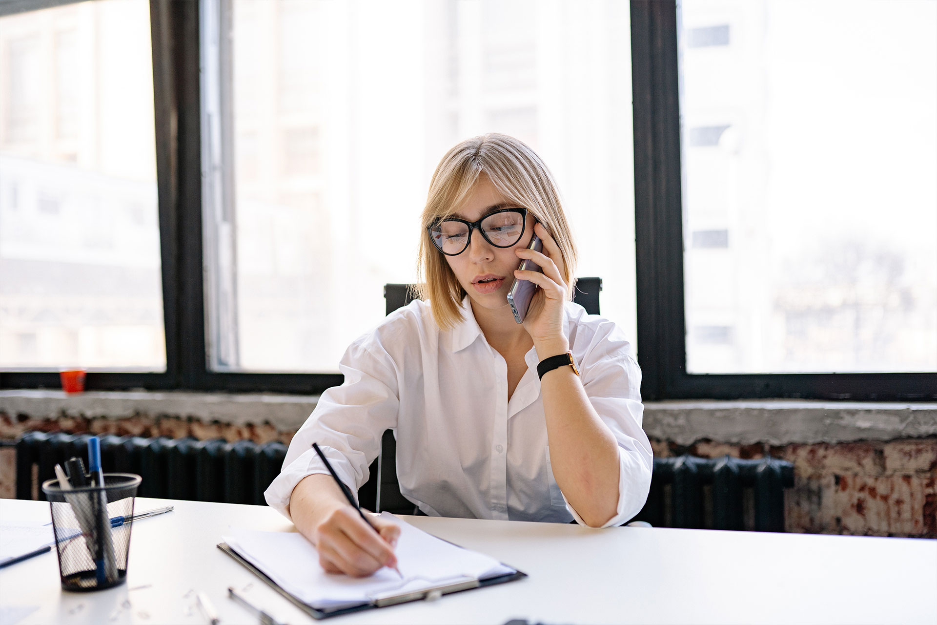 Blone Frau sitzt telephonierend am Arbeitstisch und macht Notizen