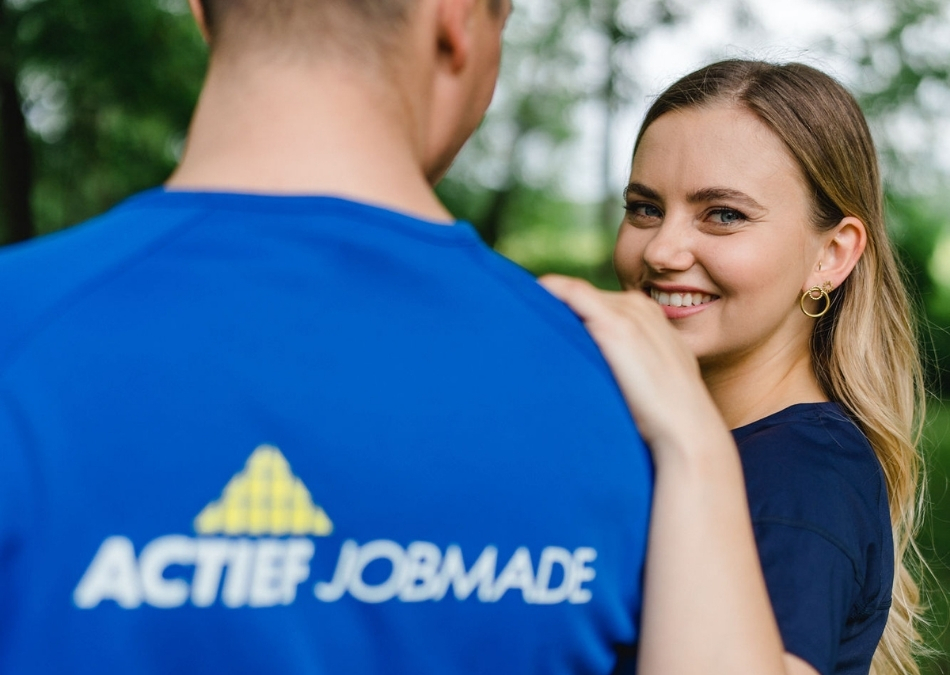 Lächelnde Frau legt ihre Hand auf die Schulter eines Mannes im blauen ACTIEF JOBMADE-Shirt im Grünen.